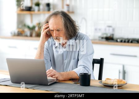 Frustré âgé caucasien gris cheveux homme, retraité, est assis à une table avec un ordinateur portable dans la cuisine, se sent surtravaillé et stressé, fatigue de travailler sur un ordinateur portable, a mal de tête, besoin de repos Banque D'Images