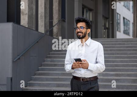 Un jeune homme, un homme d'affaires, un programmeur se promène dans la ville, tient le téléphone dans ses mains, sourit contentement, tape des messages et navigue sur Internet. Banque D'Images