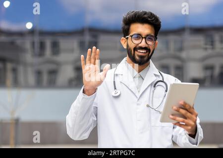 Un jeune médecin latino-américain sourit et regarde la caméra, l’homme tient une tablette dans ses mains et agite un geste amical de salutation, marche à l’extérieur de la clinique dans les airs. Banque D'Images