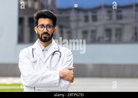 Portrait de jeune médecin confiant et sérieux, homme pensant regarder la caméra avec les bras croisés, homme indien à l'extérieur de la clinique en manteau médical blanc. Banque D'Images
