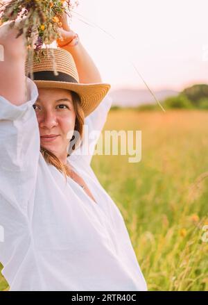 Portrait d'une jeune femme souriante vêtue de vêtements d'été légers et chapeau de paille marchant à côté d'une prairie d'herbe verte élevée avec bouquet de fleurs sauvages le soir s. Banque D'Images