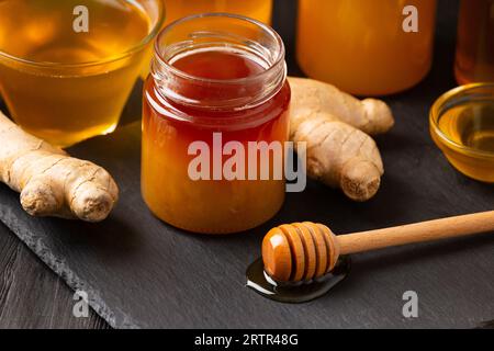 Miel dans des pots, une pelle en bois repose dans une goutte de miel, gingembre sur une assiette en ardoise en pierre noire, gros plan. Banque D'Images