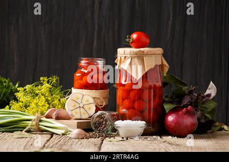 Tomates cerises en conserve dans des pots fermés et ouverts, épices et herbes pour marinade sur un fond en bois rustique. Banque D'Images