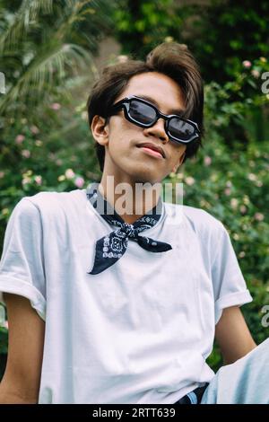 Un homme asiatique portant des lunettes de soleil et un t-shirt blanc est assis sur une table en bois dans un parc le matin Banque D'Images