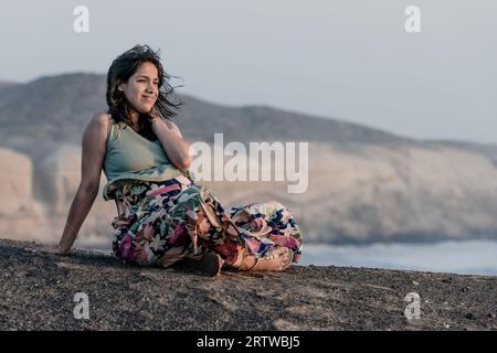 Femme enceinte assise à la plage contre le ciel pendant le coucher du soleil Banque D'Images