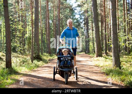 Homme athlétique en sportswear courant avec poussette de bébé au matin ensoleillé dans le parc de la ville pendant le temps de paternité. Banque D'Images