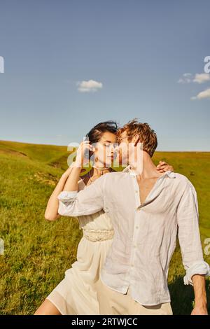 couple de jeunes mariés multiethniques en tenue de mariage blanche s'embrassant sur la prairie verte, sérénité et tendresse Banque D'Images