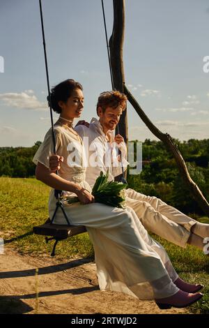 heureuse mariée asiatique avec bouquet balançant avec marié rousse dans la campagne pittoresque, mariage rustique Banque D'Images