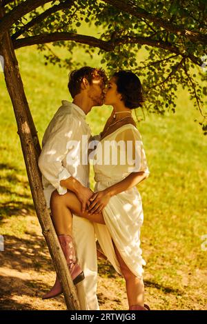 nouveaux mariés interraciaux en tenue de mariage de style boho s'embrassant sous l'arbre près de l'échelle, cadre rural Banque D'Images