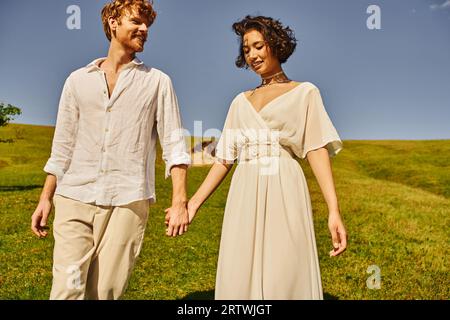 heureux moment de jeunes mariés interraciaux dans la tenue de style boho tenant les mains dans le champ, mariage rural Banque D'Images