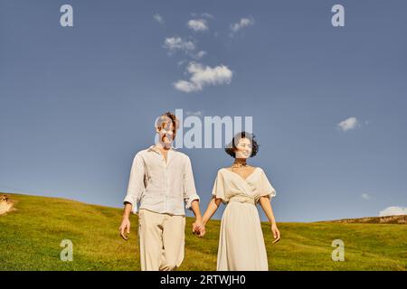 mariage rustique, joyeux couple multiethnique en tenue de style boho tenant les mains et marchant dans le pré Banque D'Images