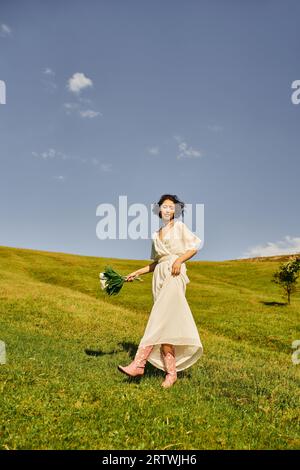 femme asiatique insouciante en robe de mariée et bottes de cow-boy marchant avec des fleurs dans le champ vert Banque D'Images