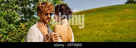 style boho, heureux couple multiethnique dans les lunettes de soleil et robe de mariée serrant dans la campagne, bannière Banque D'Images