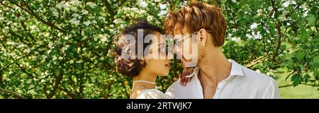 jeune couple multiethnique en lunettes de soleil et tenue blanche serrant dans la campagne, style boho, bannière Banque D'Images