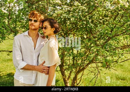 jeunes jeunes mariés multiethniques dans des lunettes de soleil et robe de mariée serrant dans le jardin vert, style boho Banque D'Images