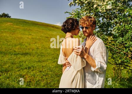 style boho, heureux jeunes mariés multiethniques dans des lunettes de soleil et robe de mariée serrant dans la campagne Banque D'Images