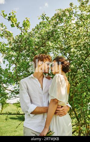 heureux jeunes mariés multiethniques dans des lunettes de soleil et robe de mariée serrant dans la campagne, style boho Banque D'Images