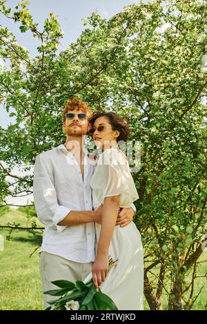 heureux jeunes mariés multiethniques dans des lunettes de soleil et robe de mariée serrant dans le jardin vert, style boho Banque D'Images