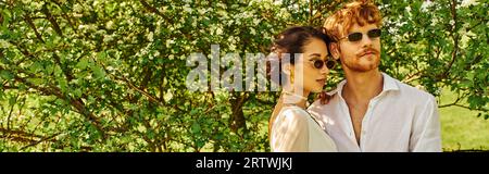 jeunes jeunes mariés multiethniques dans des lunettes de soleil et robe de mariée serrant dans le jardin vert, bannière Banque D'Images