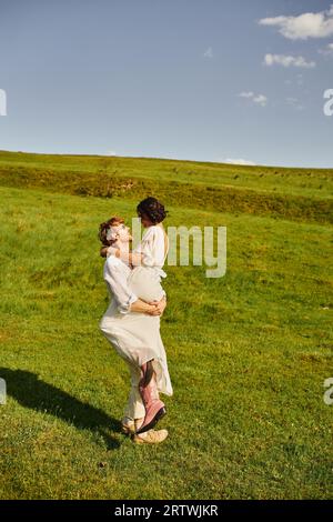 joyeux jeunes mariés, heureux marié soulevant femme asiatique en robe blanche et bottes de cow-boy dans le champ vert Banque D'Images