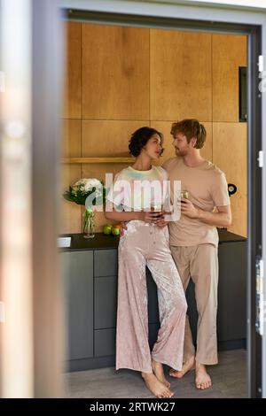 beau couple interracial debout en pyjama et tenant des tasses avec le café du matin dans la cuisine Banque D'Images