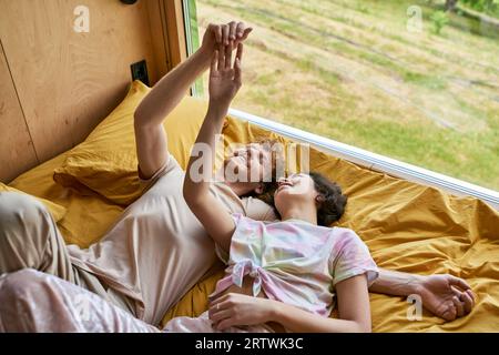 homme heureux et jeune femme asiatique touchant les mains et couchés ensemble sur la literie jaune à côté de la fenêtre Banque D'Images