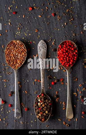 Vieilles cuillères avec graines de lin, poivre rouge et mélangé sur fond noir. Pose à plat. Vue de dessus. Concept alimentaire. Photographie culinaire d'ambiance sombre. Banque D'Images