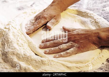 Gros plan des mains d'un pizzaiolo étirant habilement la pâte à pizza sur une surface farinée, capturant l'art de la fabrication traditionnelle de pizzas. Banque D'Images