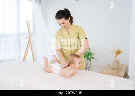 Portrait de jolie masseuse féminine faisant massage sur bébé bébé en bonne santé allongé sur la table au salon moderne. Nouveau-né allongé sur le ventre, maman aimante jouant Banque D'Images