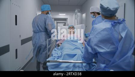 Les médecins, les infirmières et le personnel paramédical poussent la civière avec le patient malade à la salle d'opération. Femme âgée se trouve sur la civière avec goutte-à-goutte médicale. Le personnel travaille au service des urgences. Vue arrière. Tir de suivi. Banque D'Images