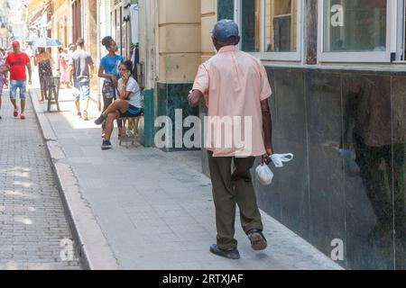 La Havane, Cuba, 2023 ans, Un cubain âgé marche sur le trottoir transportant la ration quotidienne du pain dans un sac en plastique. D'autres personnes dans la scène de la ville. Banque D'Images