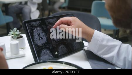 Docteur regarde l'image de l'IRM ou CT scan utilisant la tablette numérique dans le café de l'hôpital. Medic mange son dîner, analyse les résultats du scanner cérébral du patient. Le personnel médical mange à la cafétéria de la clinique. Gros plan. Banque D'Images