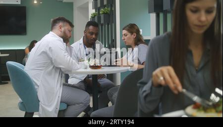 Les médecins multiethniques sont assis dans le café de la clinique, utilisent un ordinateur portable. Les médecins professionnels parlent, examinent les résultats des tests médicaux des patients, discutent du diagnostic. Le personnel médical travaille dans la cafétéria de l'hôpital ou du centre médical. Banque D'Images