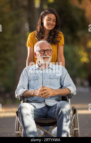 Jolie jeune femme poussant l'homme senior en fauteuil roulant dans le parc Banque D'Images