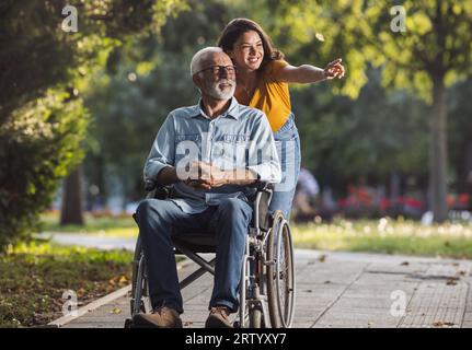 Jolie jeune femme poussant père souriant en fauteuil roulant dans le parc et ayant du bon temps à pied Banque D'Images