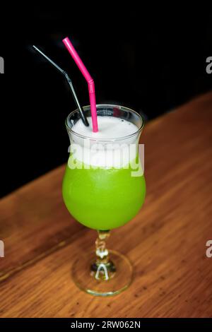 Un cocktail vert avec de la mousse blanche dans un verre avec des pailles se dresse sur un comptoir de bar en bois. Banque D'Images