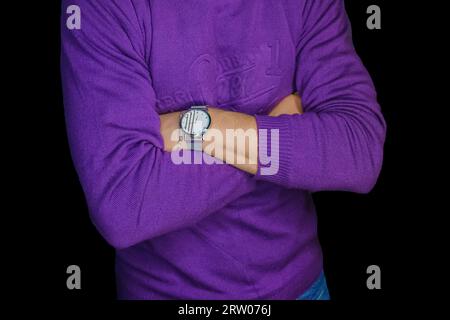 Homme posant les bras croisés avec une montre-bracelet dans un pull violet, mode et style de vêtements sur fond noir foncé. Banque D'Images