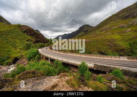 Route qui traverse la vallée de Glencoe entre hautes montagnes et prairies verdoyantes, Écosse, Royaume-Uni. Banque D'Images