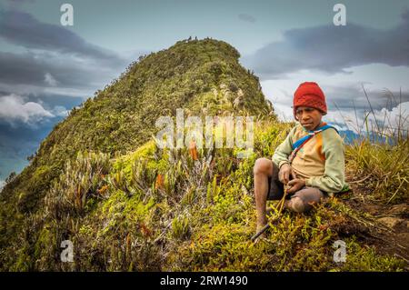 Sara village, Mt. Michael, Papouasie-Nouvelle-Guinée, juillet 2015 : un jeune garçon indigène au chapeau rouge est assis sur le sol près du sommet de la montagne et regarde tristement vers le bas Banque D'Images