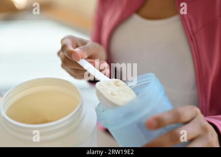 Jeune femme athlétique versant de la poudre de protéine dans une tasse, buvant un supplément pour le muscle après l'entraînement à la maison Banque D'Images