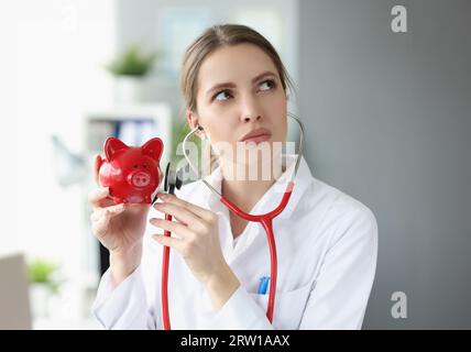 La femme médecin en couche blanche tient le stéthoscope et l'applique à la banque de porc Banque D'Images