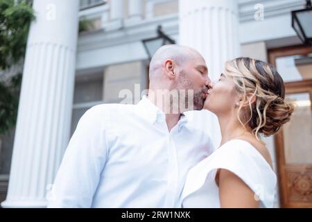 Beau marié embrasse charmante mariée sur fond de construction avec de grandes colonnes. Jeune homme en chemise blanche embrasse belle femme en élégant Banque D'Images