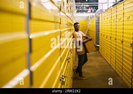 Mec souriant en jeans se tient avec une boîte en carton près des conteneurs de stockage Banque D'Images