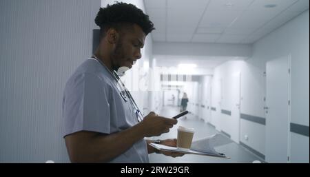 Spécialiste de la santé afro-américain avec café et dossier se tient dans le mur penché du couloir de l'hôpital et prend le téléphone. Le médecin attend rendez-vous avec le patient dans le couloir de la clinique. Le personnel médical au travail. Banque D'Images