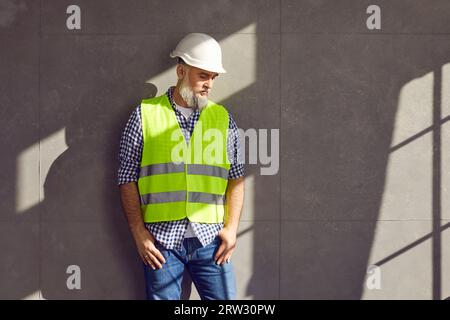 Portrait d'un contremaître barbu mature dans un casque et gilet de construction sur fond de mur gris. Banque D'Images