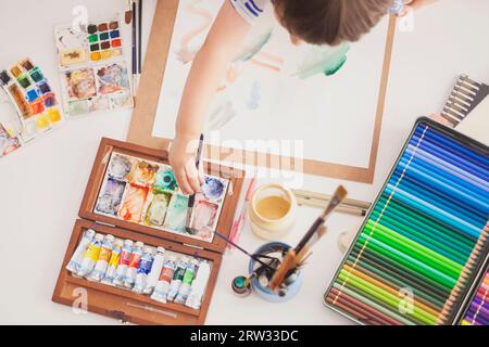 Vue de dessus d'une table avec du matériel d'illustration et les mains d'un enfant les utilisant. Banque D'Images