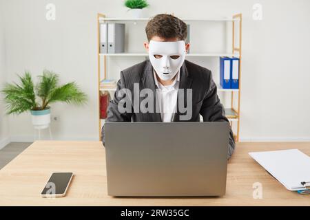 Homme d'affaires drôle portant un masque blanc assis au bureau sur son lieu de travail et travaillant sur un ordinateur portable. Banque D'Images