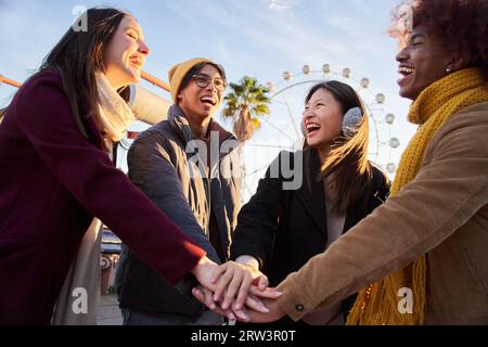Groupe de personnes multi-ethniques souriantes joignant leurs mains au parc d'attractions dans les vacances ensoleillées d'hiver. Banque D'Images