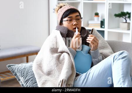 Homme asiatique malade avec des gouttes nasales et une bouteille d'eau chaude à la maison Banque D'Images