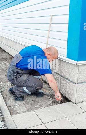 Le maçon âgé pose des tuiles sur la fondation du bâtiment à l'extérieur. Entrepreneur en construction pose des tuiles de parement de façade de l'installation de production. L'homme pose des carreaux le jour d'été. Banque D'Images